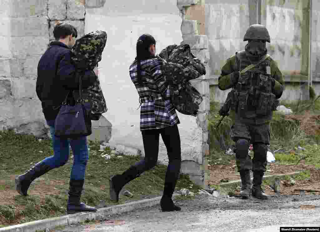 Две женщины, предположительно украинские военные, неся в руках военную форму, покидают военную базу и проходят мимо российского солдата, 19 марта 2014 года. 